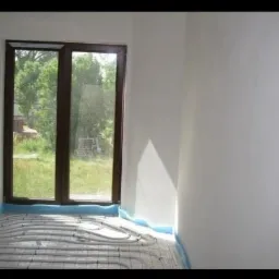 Świeżo otynkowane ściany w jasnym pomieszczeniu z oknem, widoczne rury ogrzewania podłogowego zabezpieczone niebieską folią.