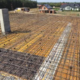 Zbrojenie płyty fundamentowej z widocznymi dystansami z tworzywa sztucznego, na deskowaniu z żółtej sklejki, z betonowym słupem w tle i zabudową jednorodzinną w oddali.