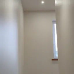 Jasne, świeżo pomalowane wnętrze z oknem i subtelnym oświetleniem punktowym. Widoczne detale wykończeniowe i neutralna kolorystyka ścian.