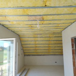 Remonty i wykończenia - Żółta wełna mineralna między metalowymi profilami na poddaszu, widok z dołu. Fragment ściany z oknem i drabina w surowym wnętrzu.
