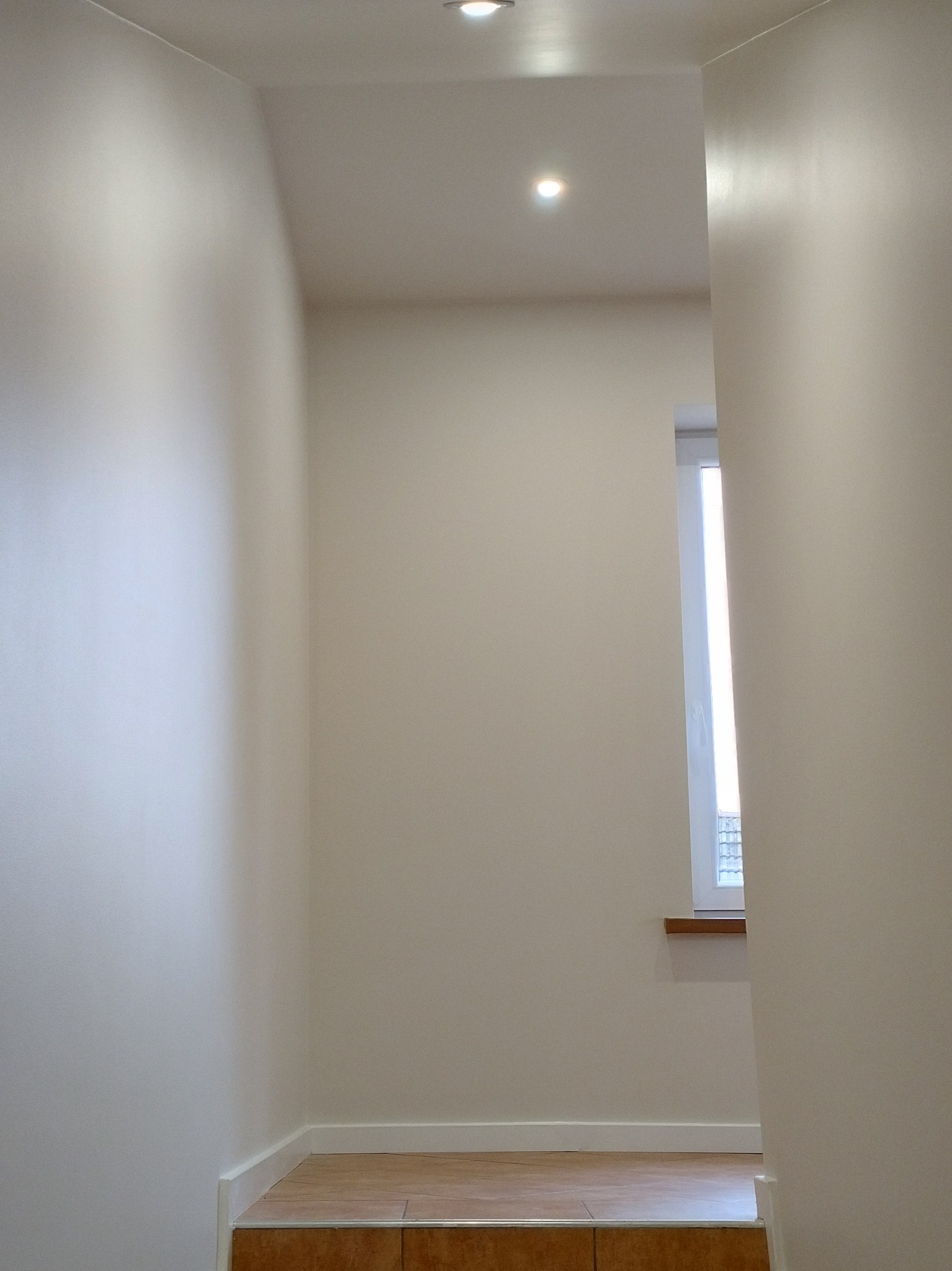 Jasne, świeżo pomalowane wnętrze z oknem i subtelnym oświetleniem punktowym. Widoczne detale wykończeniowe i neutralna kolorystyka ścian.