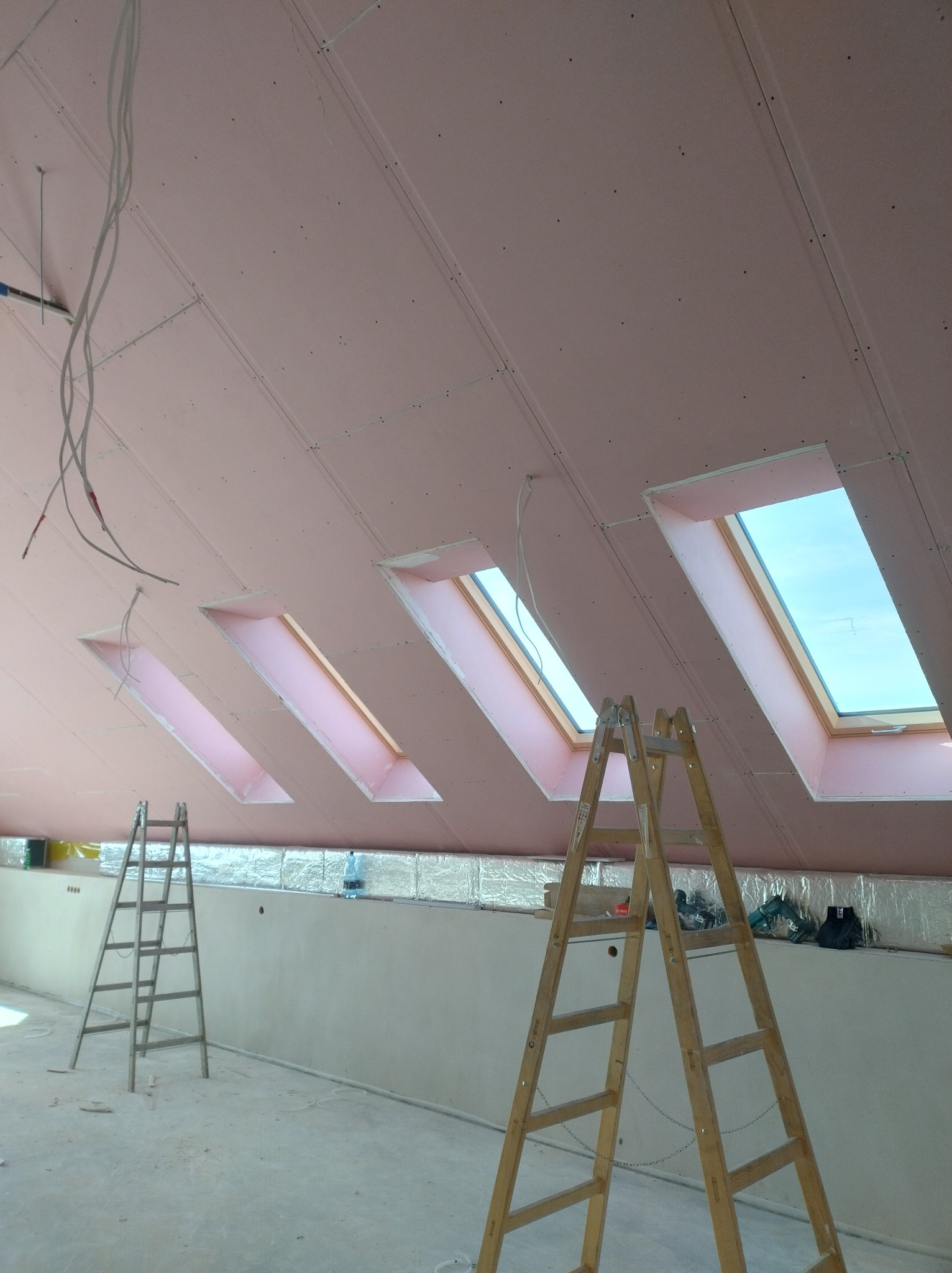 Poddasze z oknami dachowymi w trakcie wykańczania płytami gipsowo-kartonowymi. Widoczne dwie drabiny i izolacja. Przewody elektryczne zwisają z sufitu.