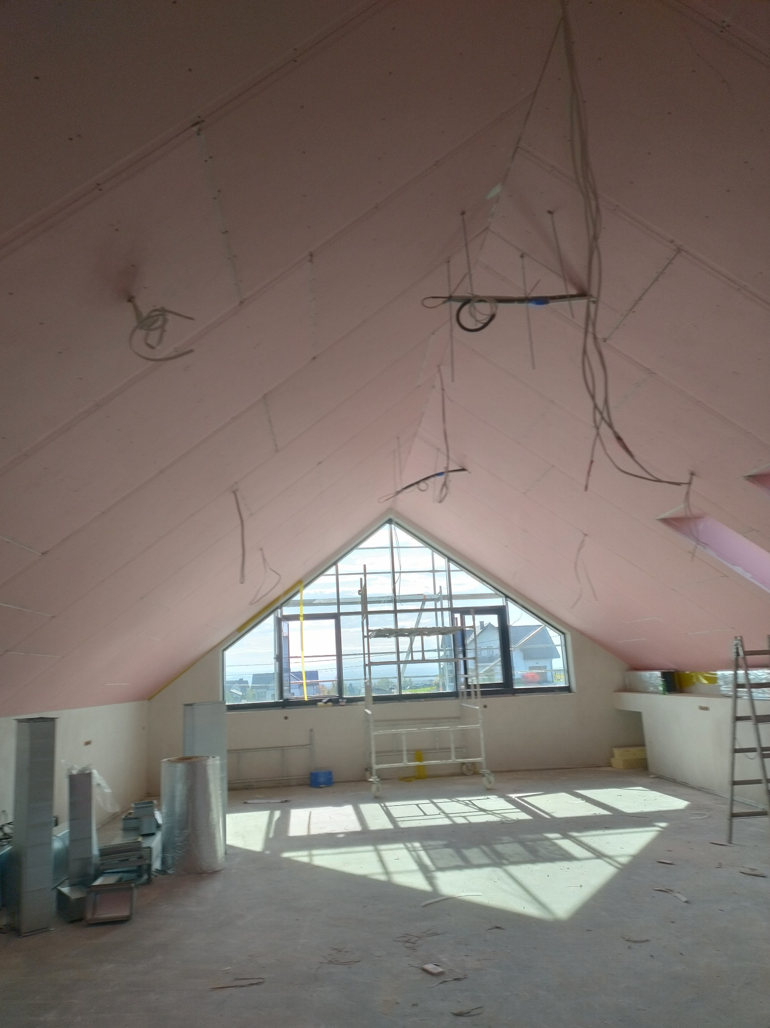 Poddasze w trakcie prac wykończeniowych: widoczne płyty karton-gips na skosach, okno dachowe, rusztowanie i elementy instalacji elektrycznej. Jasne wnętrze w surowym stanie.