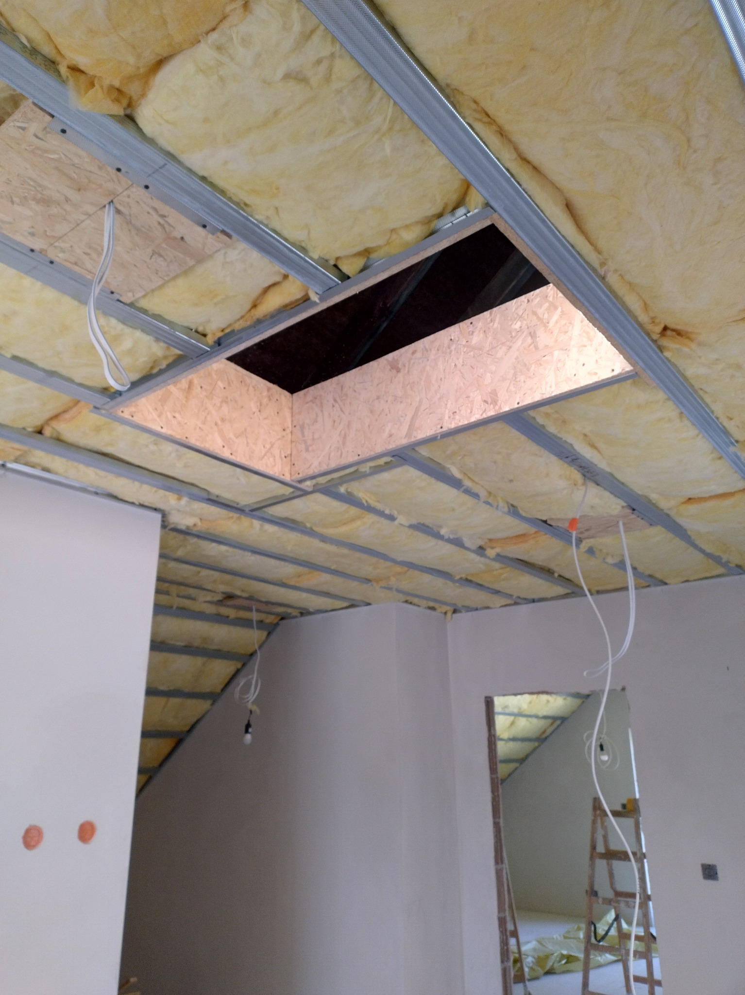 Ocieplenie poddasza wełną mineralną, widoczne metalowe profile i płyty OSB. Otwór rewizyjny w suficie. Przewody elektryczne zwisają z sufitu.