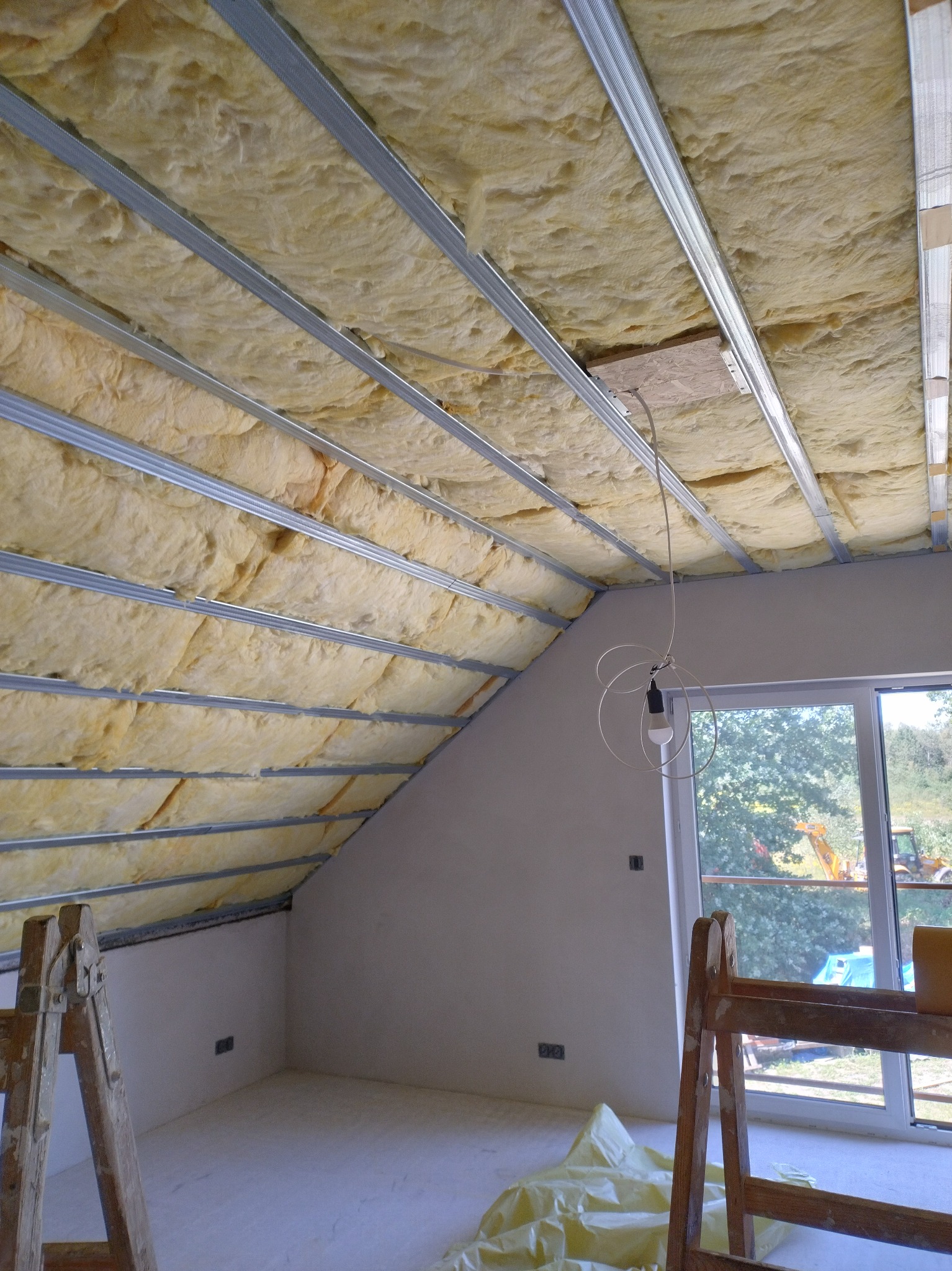 Ocieplone wełną poddasze z metalowym stelażem, widok z dołu. Fragment drabiny malarskiej w rogu kadru. Okno z widokiem na zewnątrz.