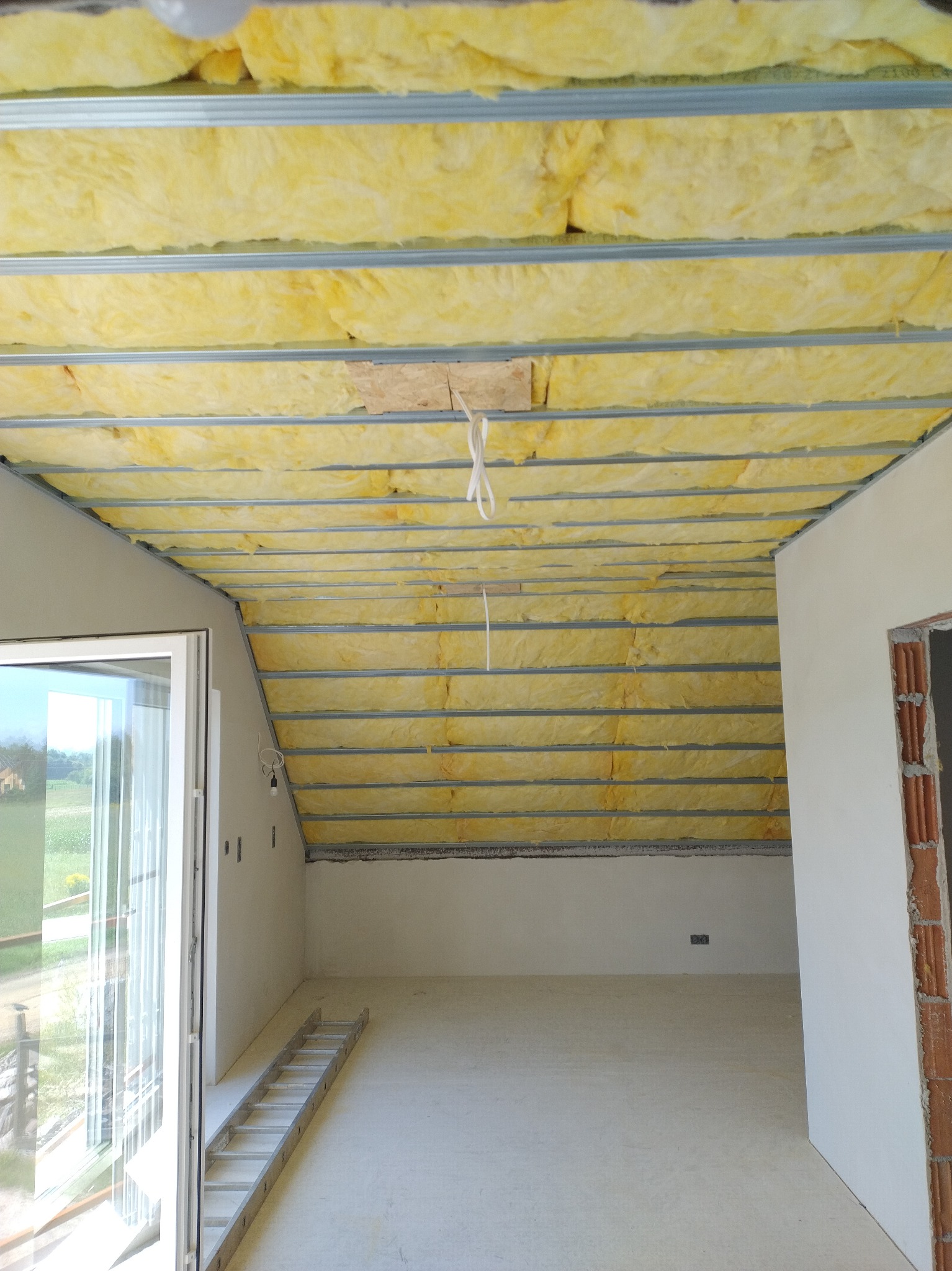 Żółta wełna mineralna między metalowymi profilami na poddaszu, widok z dołu. Fragment ściany z oknem i drabina w surowym wnętrzu.