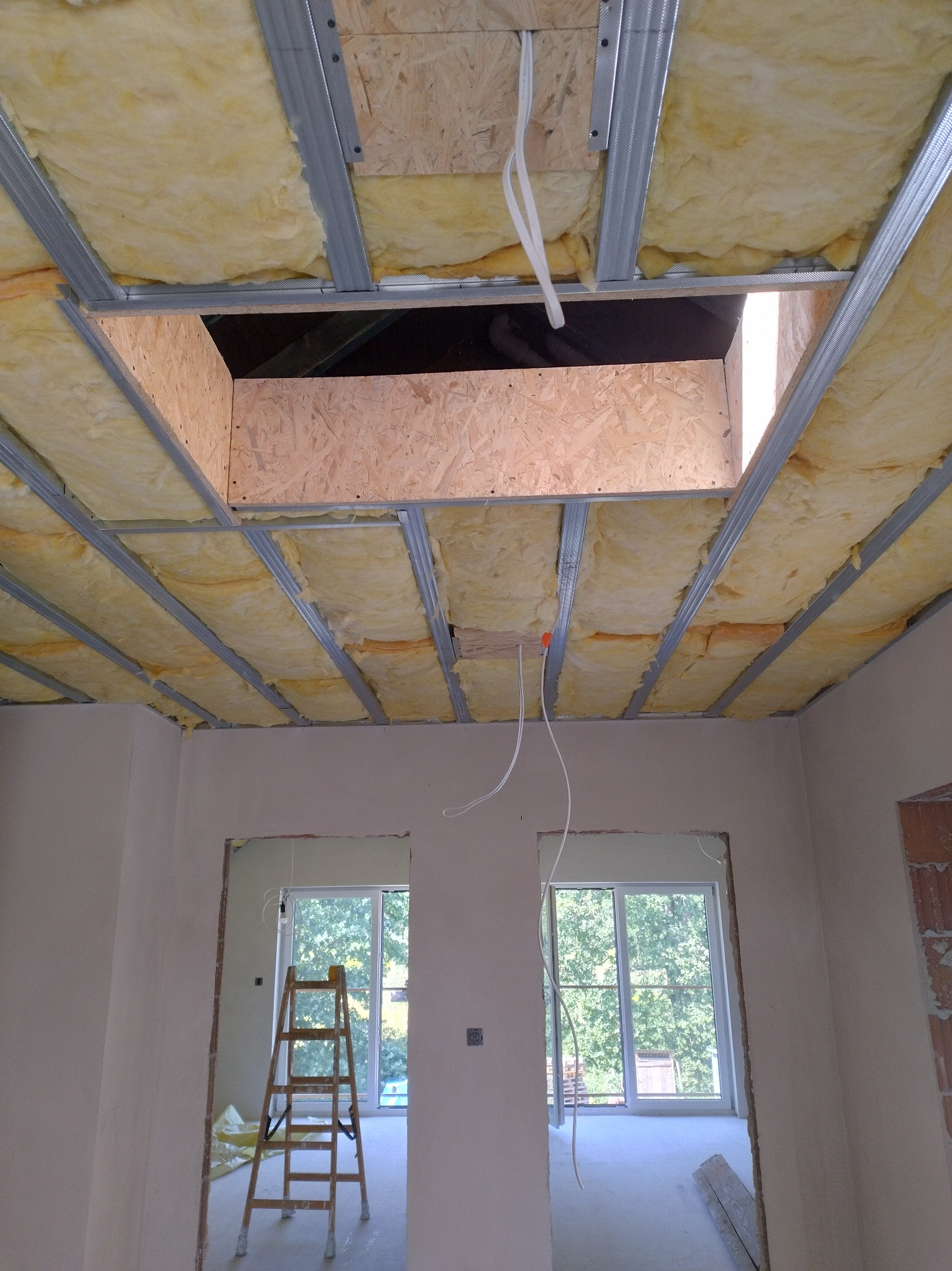 Ocieplenie poddasza wełną mineralną, widoczne metalowe profile i płyty OSB. W tle okno i drabina. Prace wykończeniowe w jasnym pomieszczeniu.