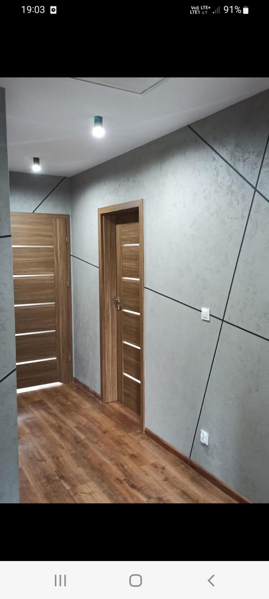 Korytarz w stylu loft z drewnianą podłogą, dwiema drewnianymi drzwiami z poziomymi panelami i szarą ścianą z geometrycznymi czarnymi liniami.