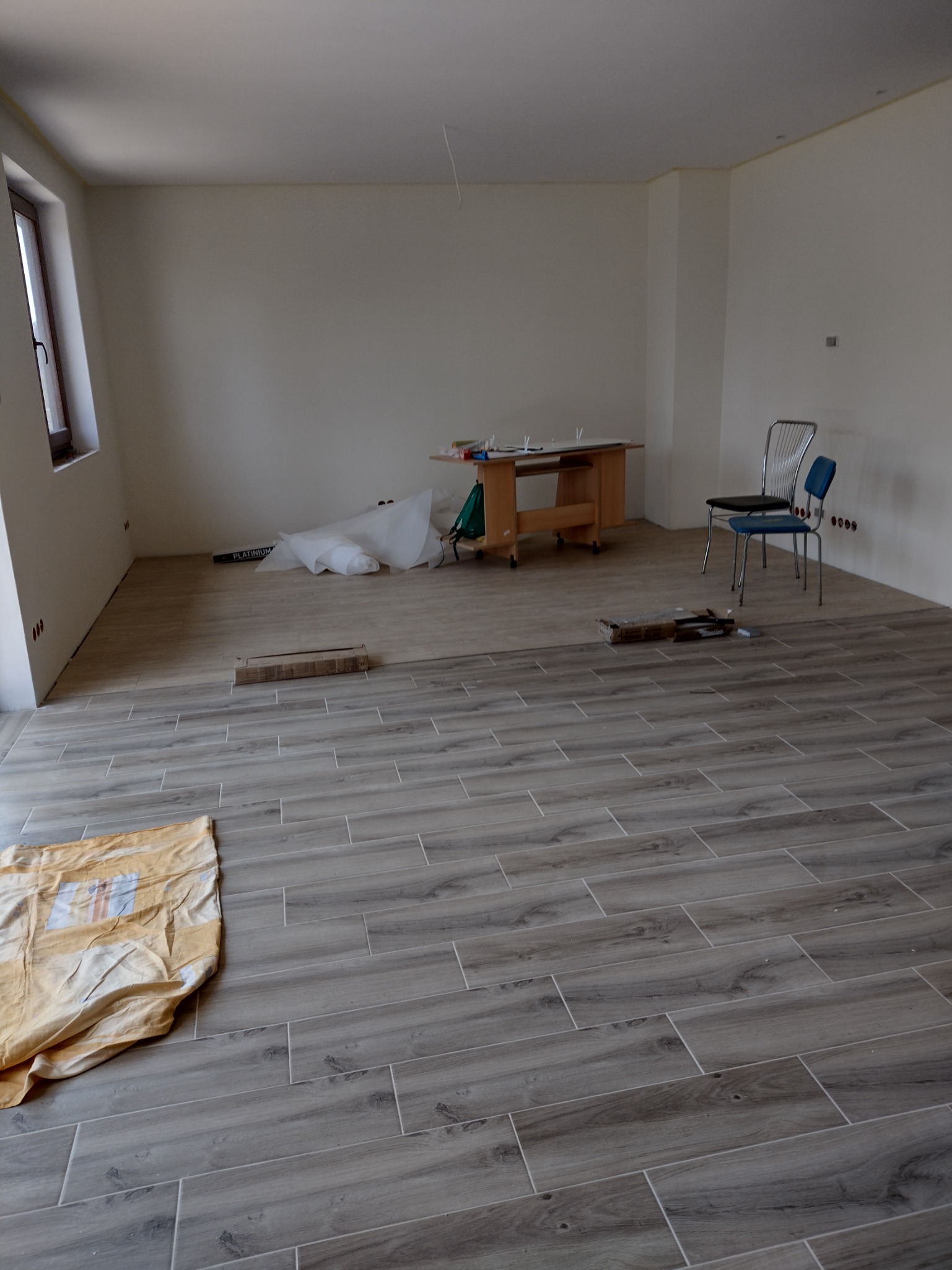 Ułożona podłoga z płytek imitujących drewno w pustym, jasnym pokoju po remoncie, widoczna plandeka ochronna, karton po płytkach i stolik roboczy.