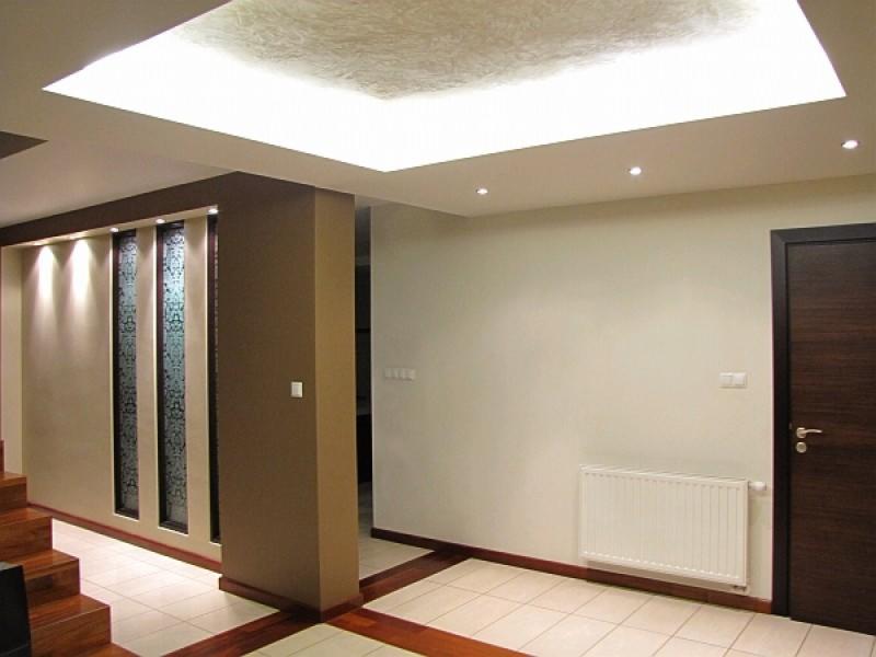 Nowoczesne wnętrze: kremowe ściany, podłoga z białych płytek i drewnianymi akcentami, drewniane schody, dekoracyjna ściana z podświetlanymi, wzorzystymi wstawkami, biały grzejnik pod ścianą, ciemne...