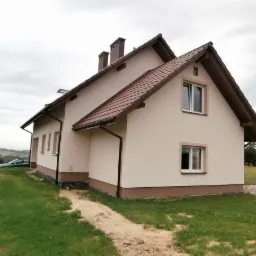 Nowo wybudowany, otynkowany dom jednorodzinny z brązową dachówką i elementami wykończeniowymi, na trawiastym terenie z widocznym samochodem w tle.