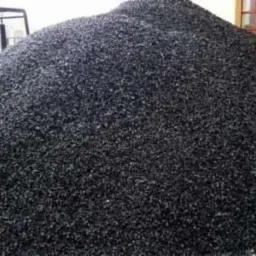 Góra czarnego, błyszczącego paliwa stałego na składzie, z widocznym w tle wózkiem widłowym i oknem.