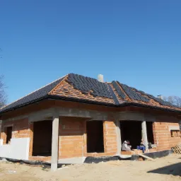 Budowa domu jednorodzinnego w stanie surowym otwartym: częściowo ułożone czarne dachówki na drewnianej konstrukcji, ściany z czerwonej cegły, ocieplenie fragmentu ściany styropianem, dwóch...