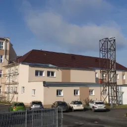 Budynek z brązowym dachem z dachówki, widoczne rusztowania i zaparkowane samochody, błękitne niebo z chmurami w tle.