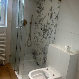 Merbud - Nowoczesna łazienka z kabiną prysznicową ze złotymi elementami, toaletą podwieszaną i płytkami z motywem kwiatowym w odcieniach szarości, drewnopodobna podłoga.