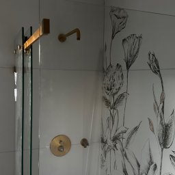 Merbud - Szklana kabina prysznicowa z złotymi detalami, białe płytki z motywem kwiatowym, nowoczesna bateria prysznicowa.