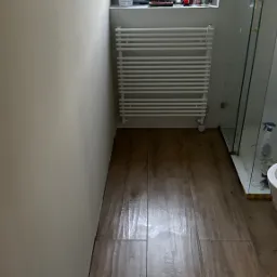 Wykończona łazienka z drewnopodobną podłogą, grzejnikiem drabinkowym pod oknem i kabiną prysznicową z przezroczystego szkła.