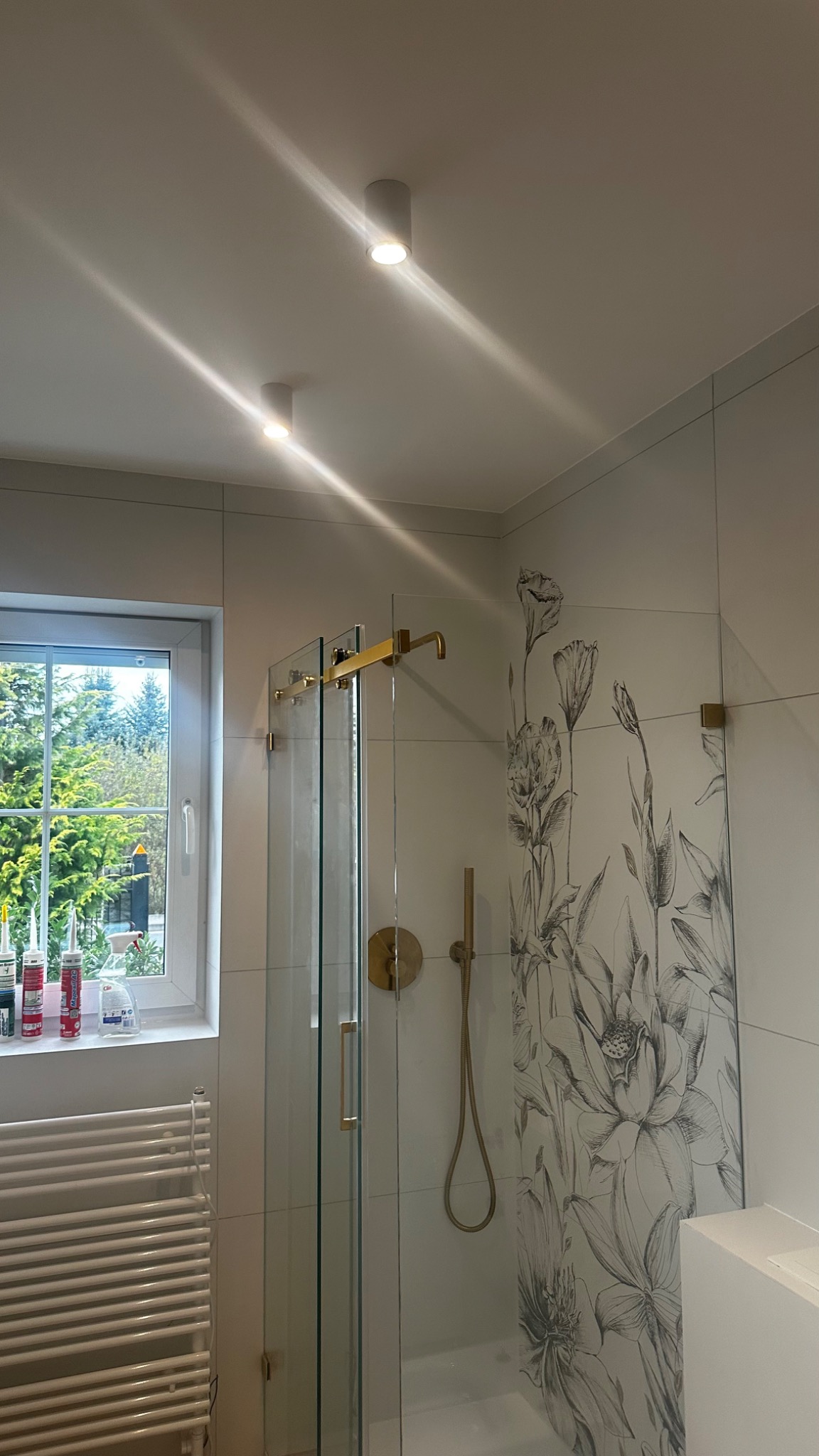 Nowoczesna kabina prysznicowa z złotymi elementami, zdobiona wzorem kwiatowym na płytkach, widok z okna na zieleń, widoczne tuby silikonu na parapecie.