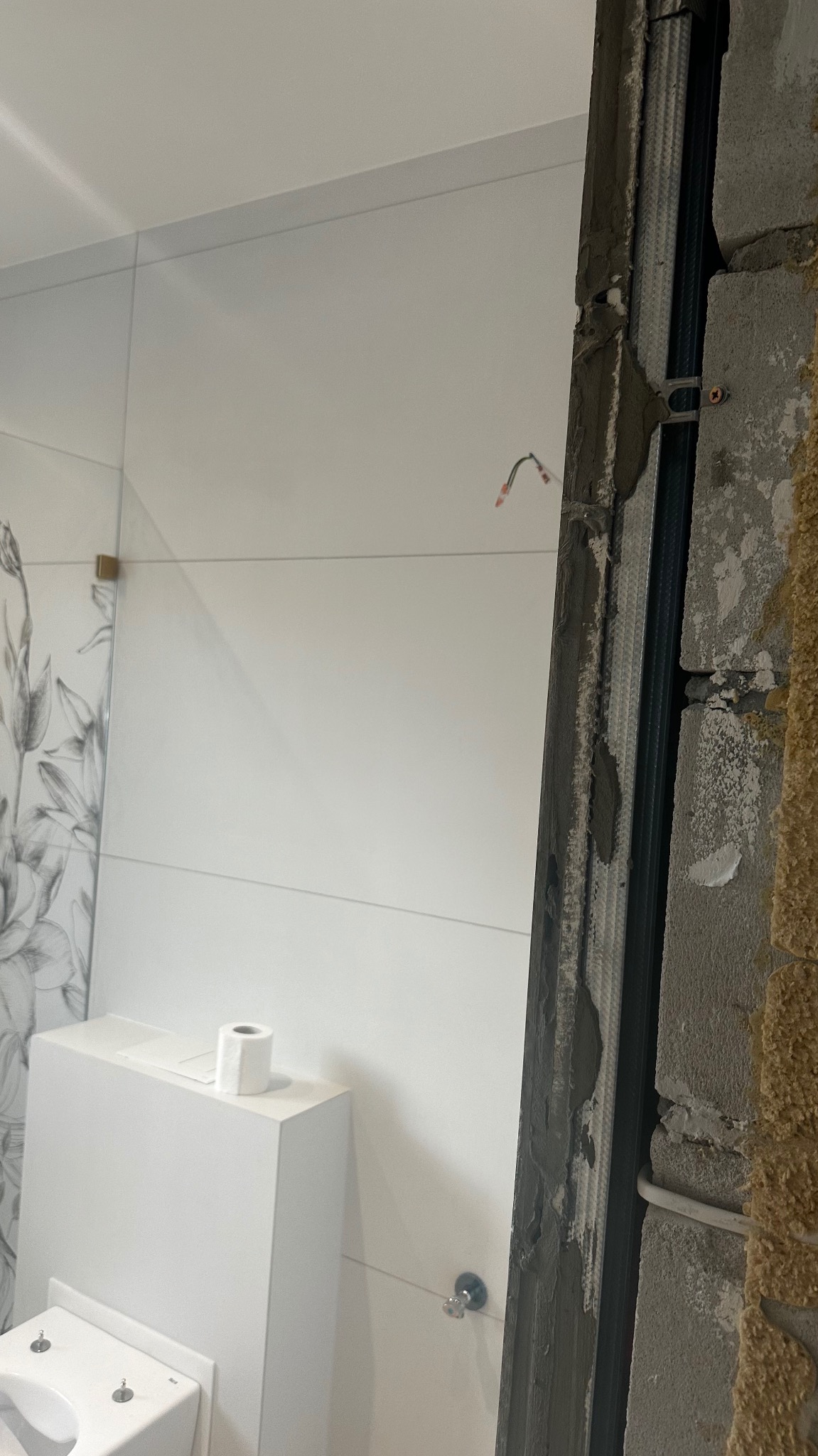 Wnętrze łazienki w trakcie remontu, widoczna zabudowa geberit z rolką papieru toaletowego, fragment ściany z płytkami z motywem roślinnym oraz fragment ściany z profilem do montażu płyty karton-gips.