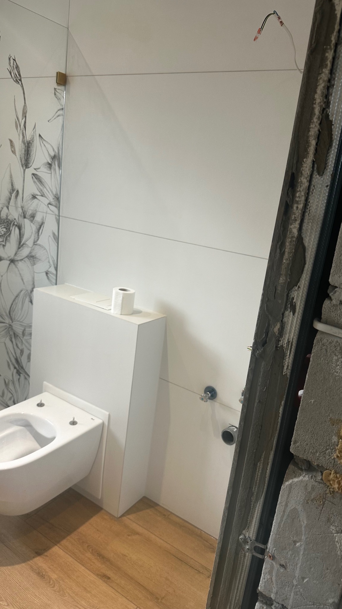 Podtynkowa toaleta w trakcie montażu, widoczne przyłącza hydrauliczne, płytki z motywem kwiatowym i surowy fragment ściany z profilem.
