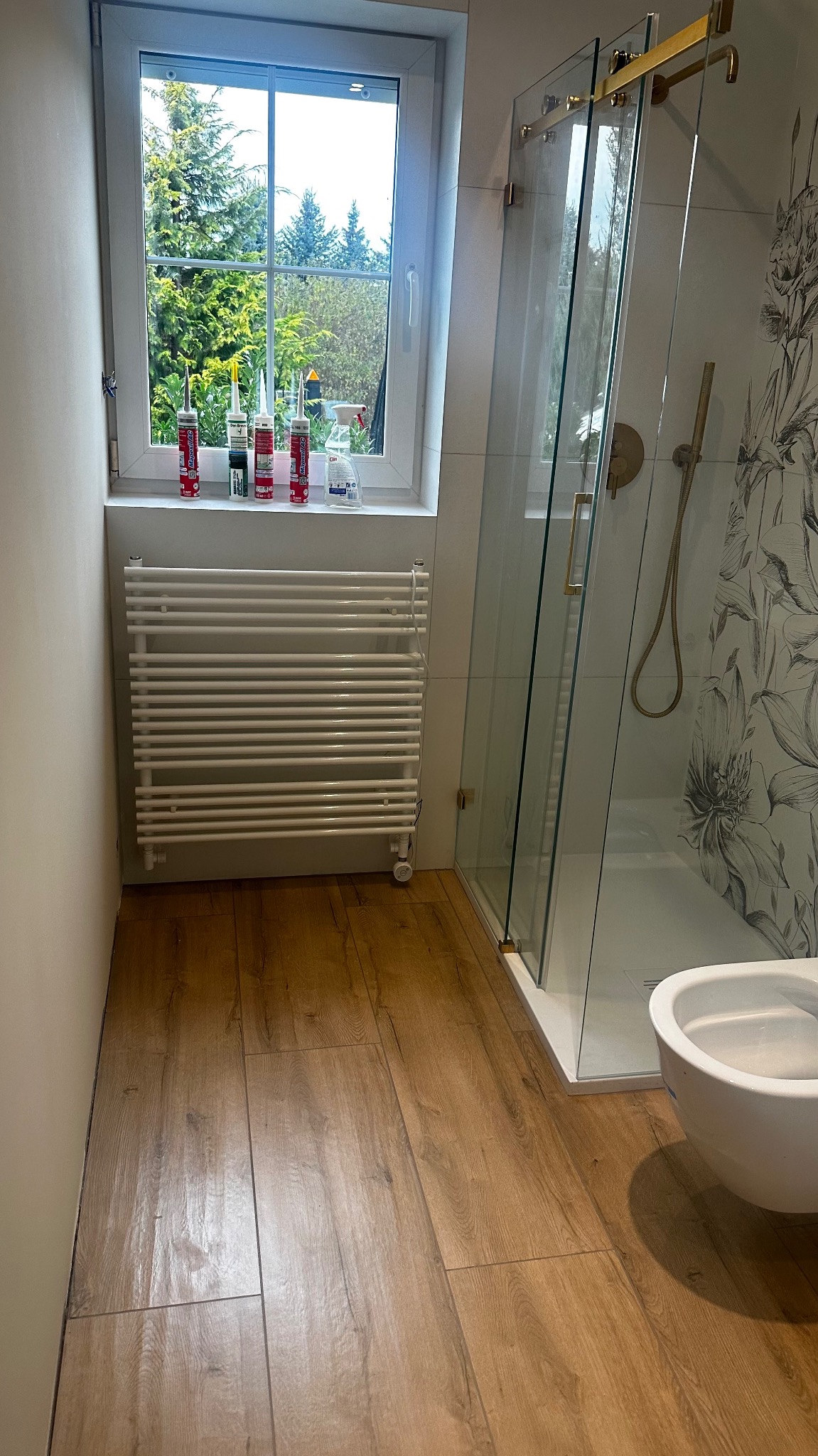 Nowoczesna łazienka z drewnopodobną podłogą, kabiną prysznicową z złotymi elementami i wiszącą toaletą. Na parapecie okna stoją silikony budowlane.