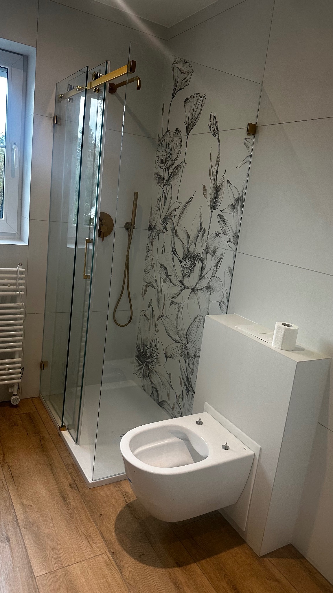 Nowoczesna łazienka z kabiną prysznicową ze złotymi elementami, toaletą podwieszaną i płytkami z motywem kwiatowym w odcieniach szarości, drewnopodobna podłoga.