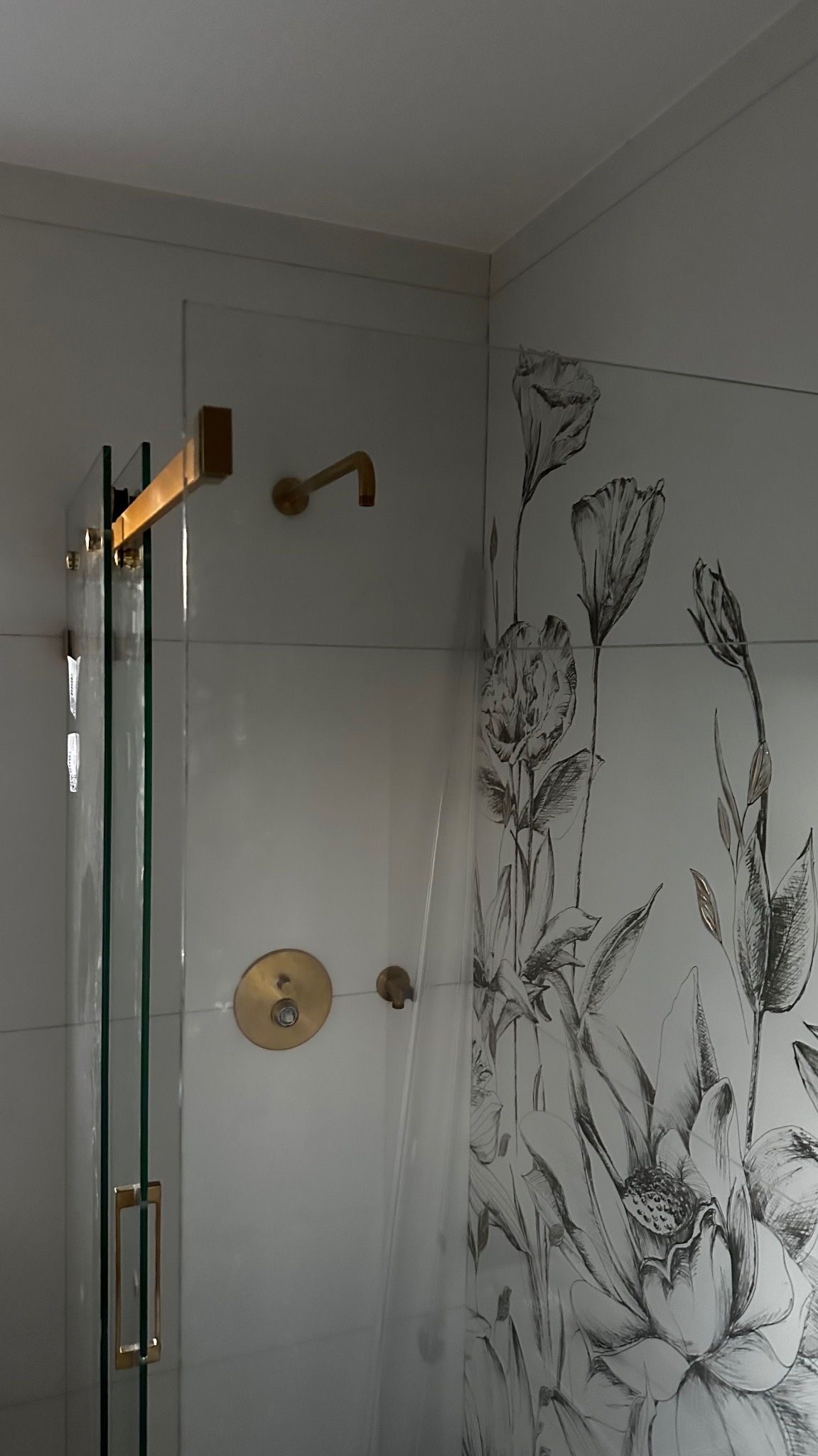 Szklana kabina prysznicowa z złotymi detalami, białe płytki z motywem kwiatowym, nowoczesna bateria prysznicowa.