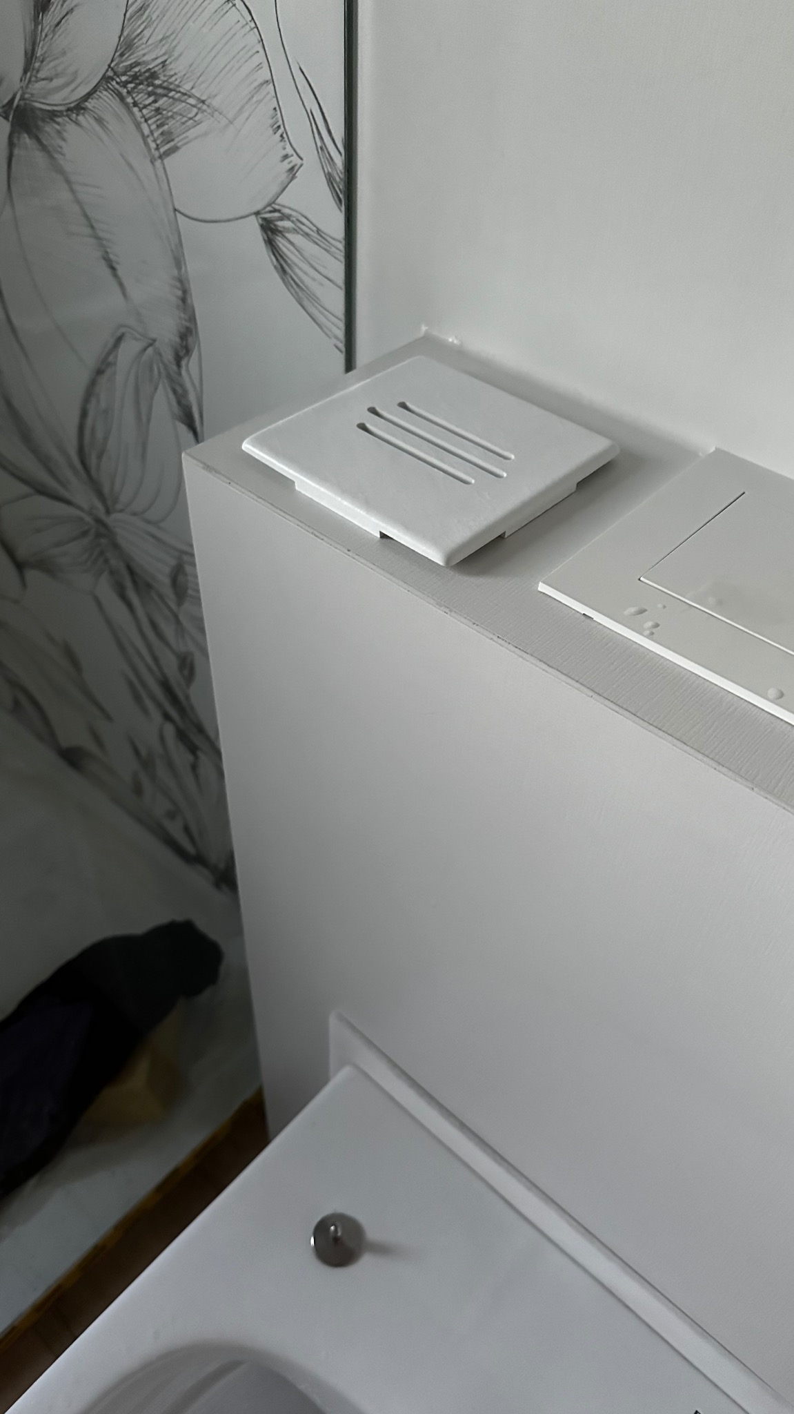 Biała zabudowa toalety z ukrytym systemem spłukiwania, widoczny fragment muszli klozetowej, dekoracyjna tapeta z motywem kwiatowym w tle.