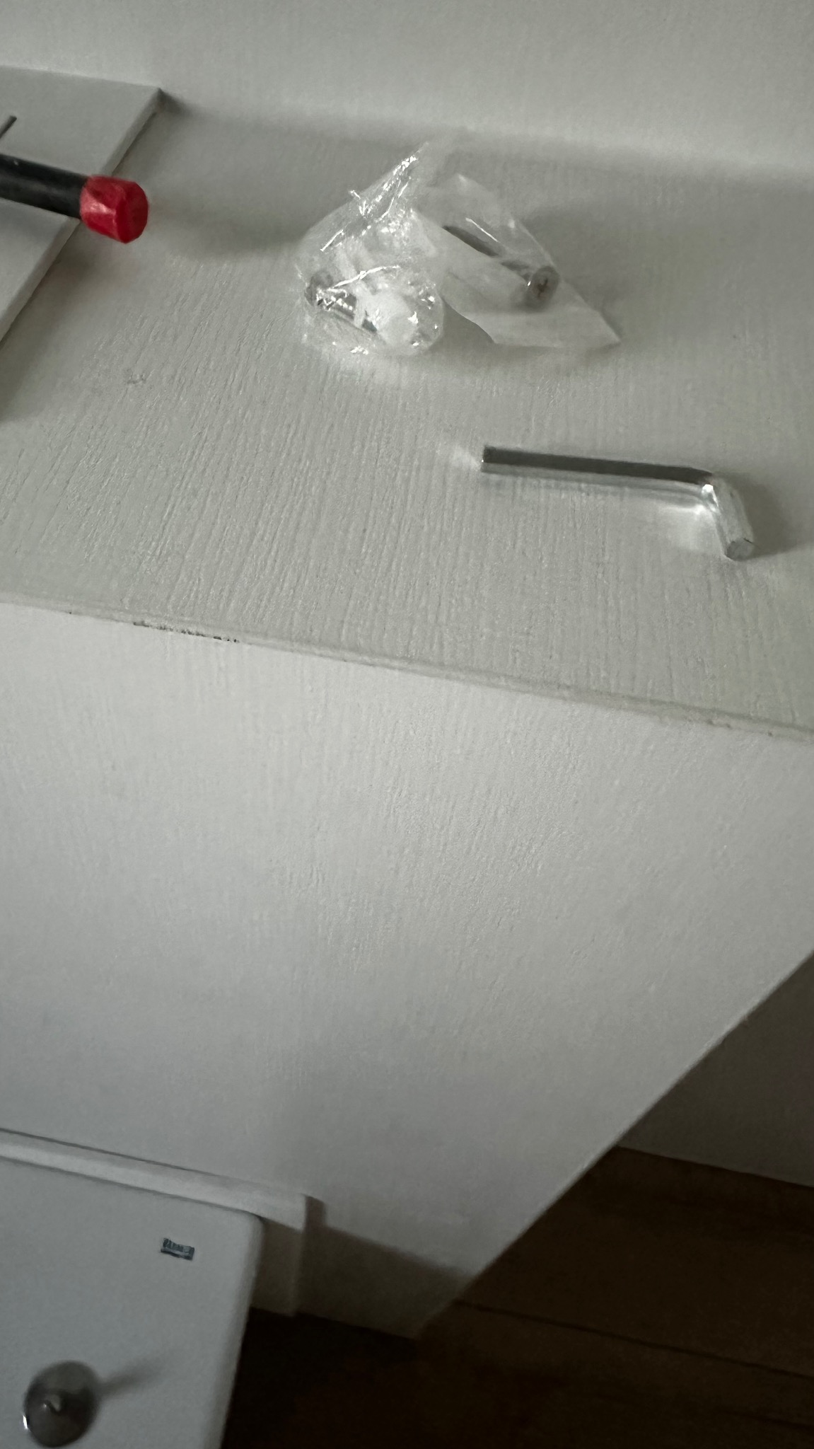 Akcesoria montażowe do mebli: klucz imbusowy, wkręty w folii, śrubokręt z czerwoną rączką leżą na białym blacie szafki.