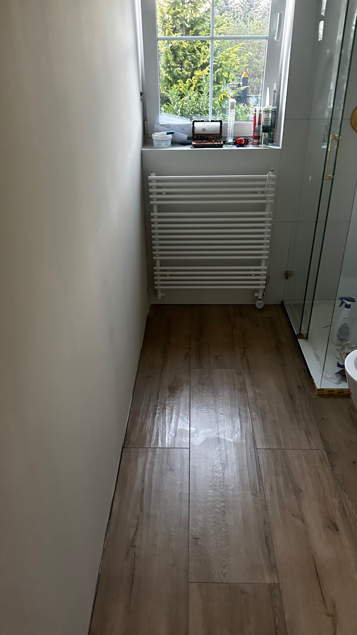 Wykończona łazienka z drewnopodobną podłogą, grzejnikiem drabinkowym pod oknem i kabiną prysznicową z przezroczystego szkła.