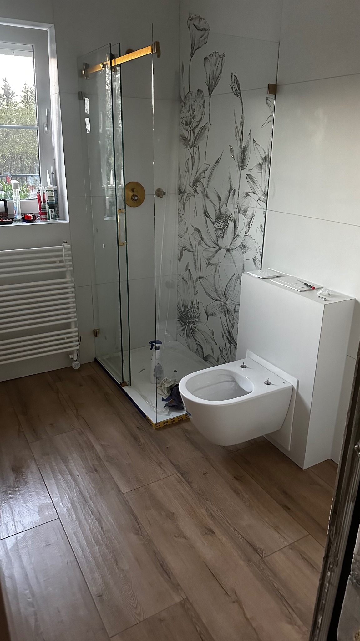 Nowoczesna łazienka w trakcie montażu: biała wisząca toaleta z zabudową, kabina prysznicowa z złotymi elementami, płytki z motywem kwiatowym, grzejnik drabinkowy i panele podłogowe imitujące drewno.