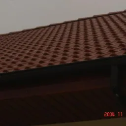 Czerwona dachówka ceramiczna ułożona na połaci dachowej, widoczne czarne rynny i podbitka dachowa z brązowego drewna, pochmurne niebo w tle.