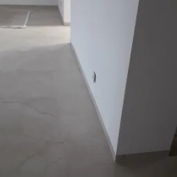 Wykończone wnętrze w Cotoń z beżową podłogą z płytek imitujących kamień i świeżo pomalowanymi białymi ścianami z widocznym gniazdkiem elektrycznym, kabel na podłodze.