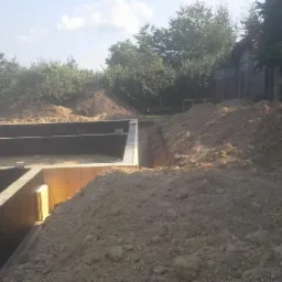 Zabezpieczone, wykopane fundamenty domu z widocznymi deskami szalunkowymi, na tle wzgórza ziemi i zieleni. Kąt ujęcia z dołu podkreśla głębokość wykopu.