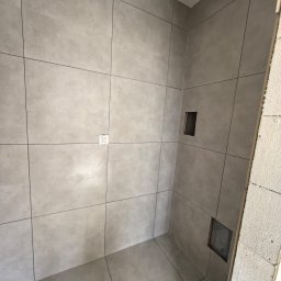 Wykończona łazienka z szarymi, matowymi płytkami wielkoformatowymi, czarnymi fugami i gniazdkiem elektrycznym. Widoczny fragment surowej ściany z bloczków.