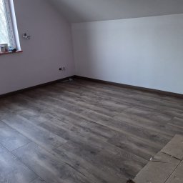 GREEN-BUD REMONTY - Ukończona podłoga z paneli w pokoju na poddaszu, widoczne fragmenty kartonów zabezpieczających podłogę. Ściany pomalowane na biało i delikatny róż.
