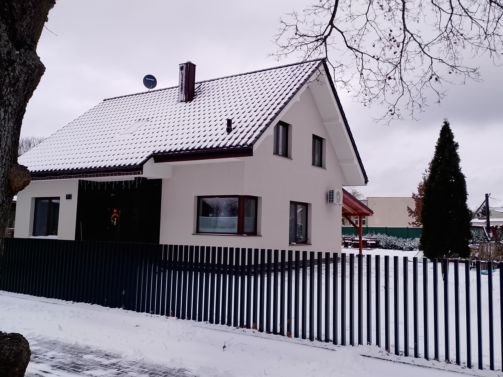 Biały dom jednorodzinny z ciemną stolarką okienną i dachem pokrytym śniegiem, widoczny zza czarnego, metalowego ogrodzenia w zimowej scenerii.
