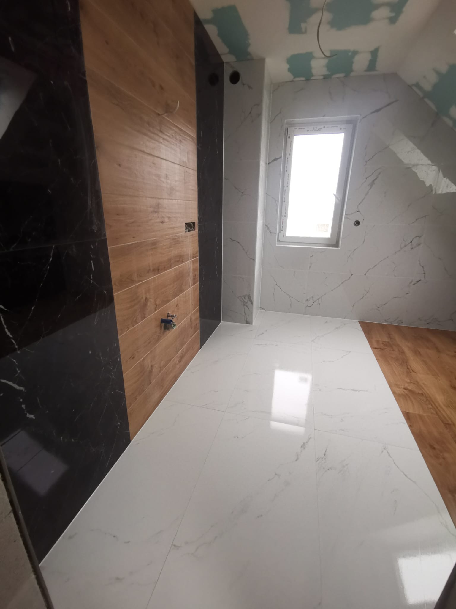 Wnętrze łazienki w trakcie wykańczania: białe marmurowe płytki na podłodze i ścianach, drewnopodobne panele na ścianie, okno w skosie, widoczne ślady prac wykończeniowych.
