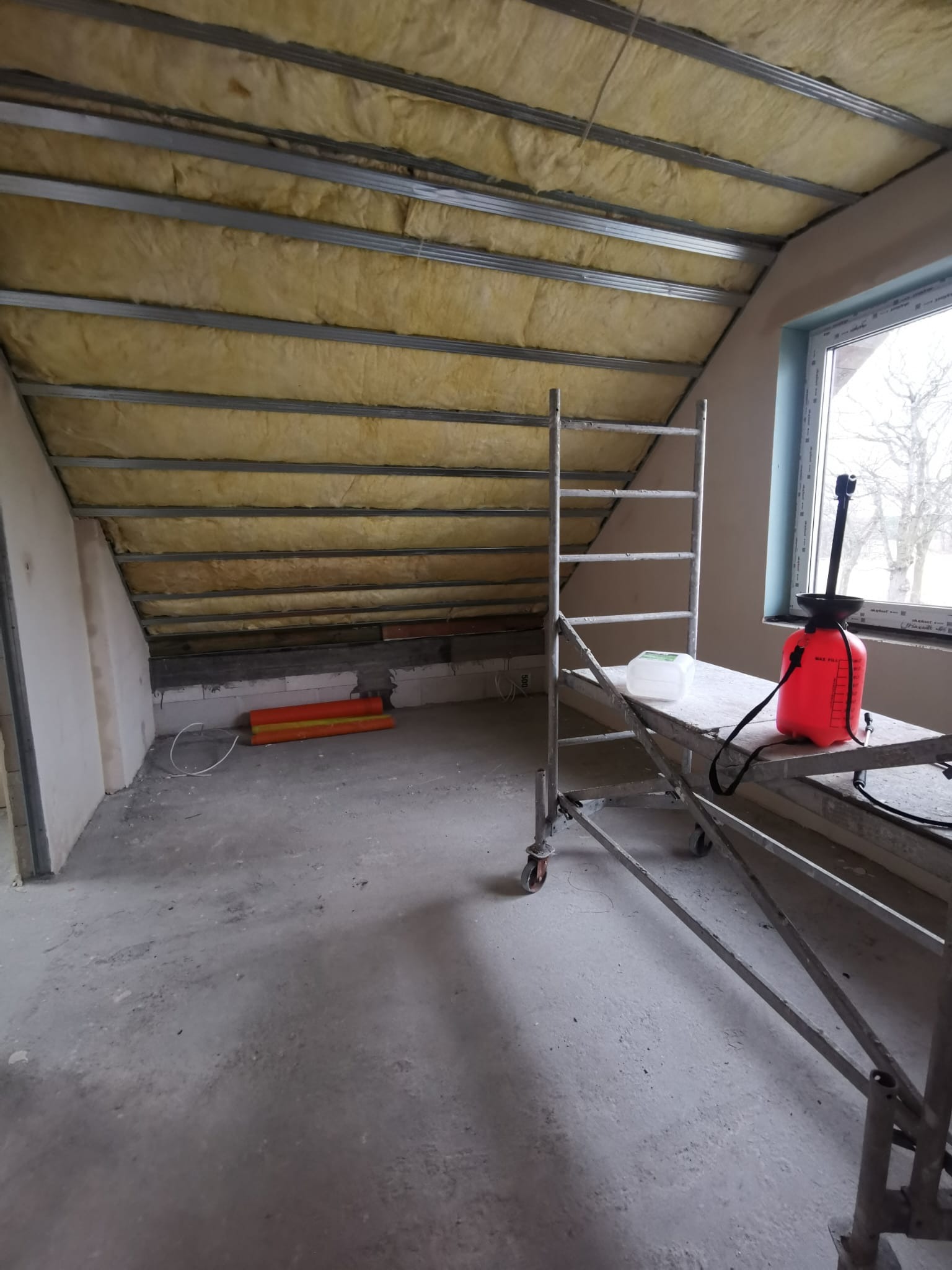 Poddasze w trakcie ocieplania wełną mineralną, widoczne metalowe profile, rusztowanie z czerwonym opryskiwaczem. Prace wykończeniowe w surowym wnętrzu.