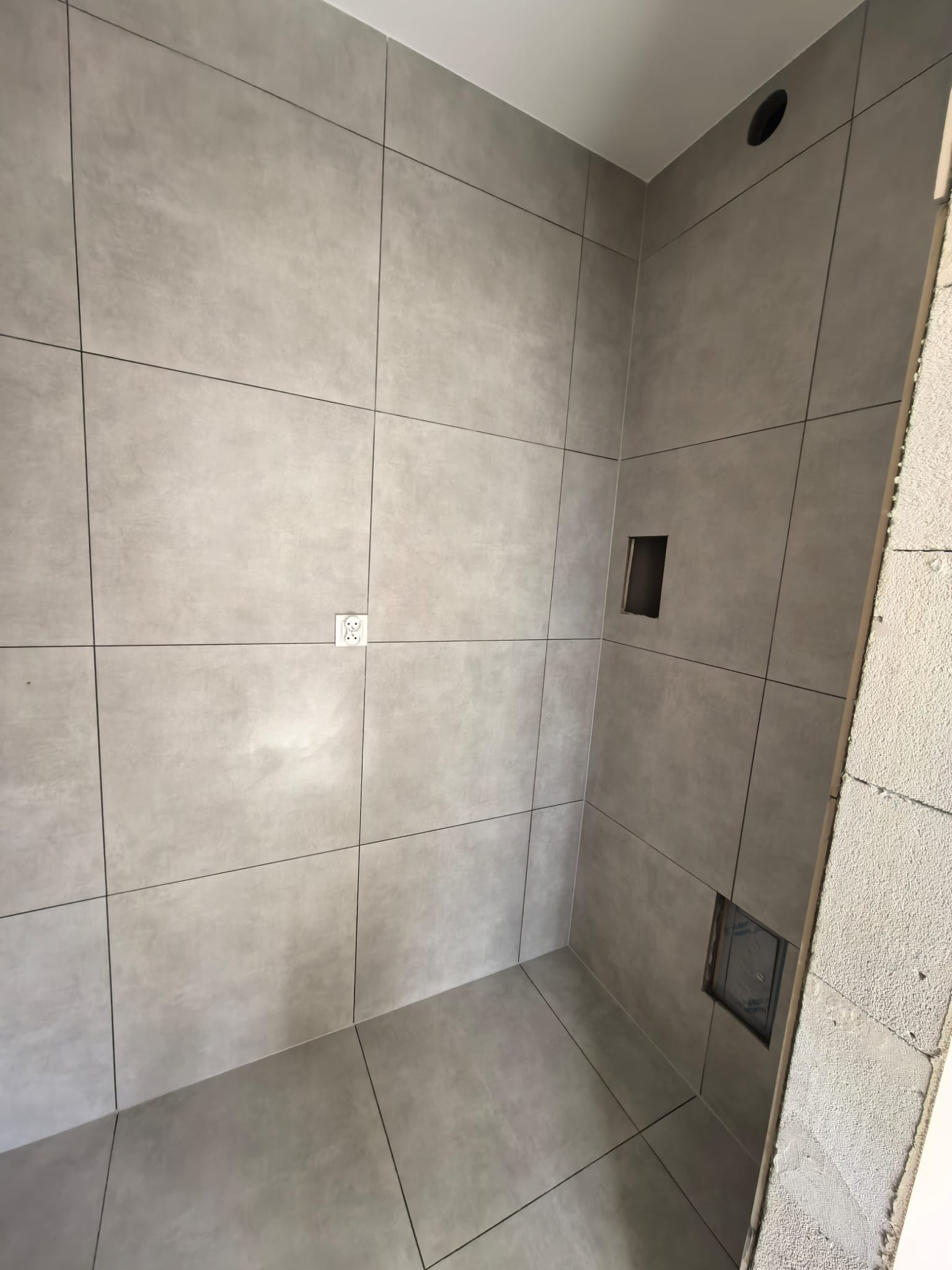 Wykończona łazienka z szarymi, matowymi płytkami wielkoformatowymi, czarnymi fugami i gniazdkiem elektrycznym. Widoczny fragment surowej ściany z bloczków.