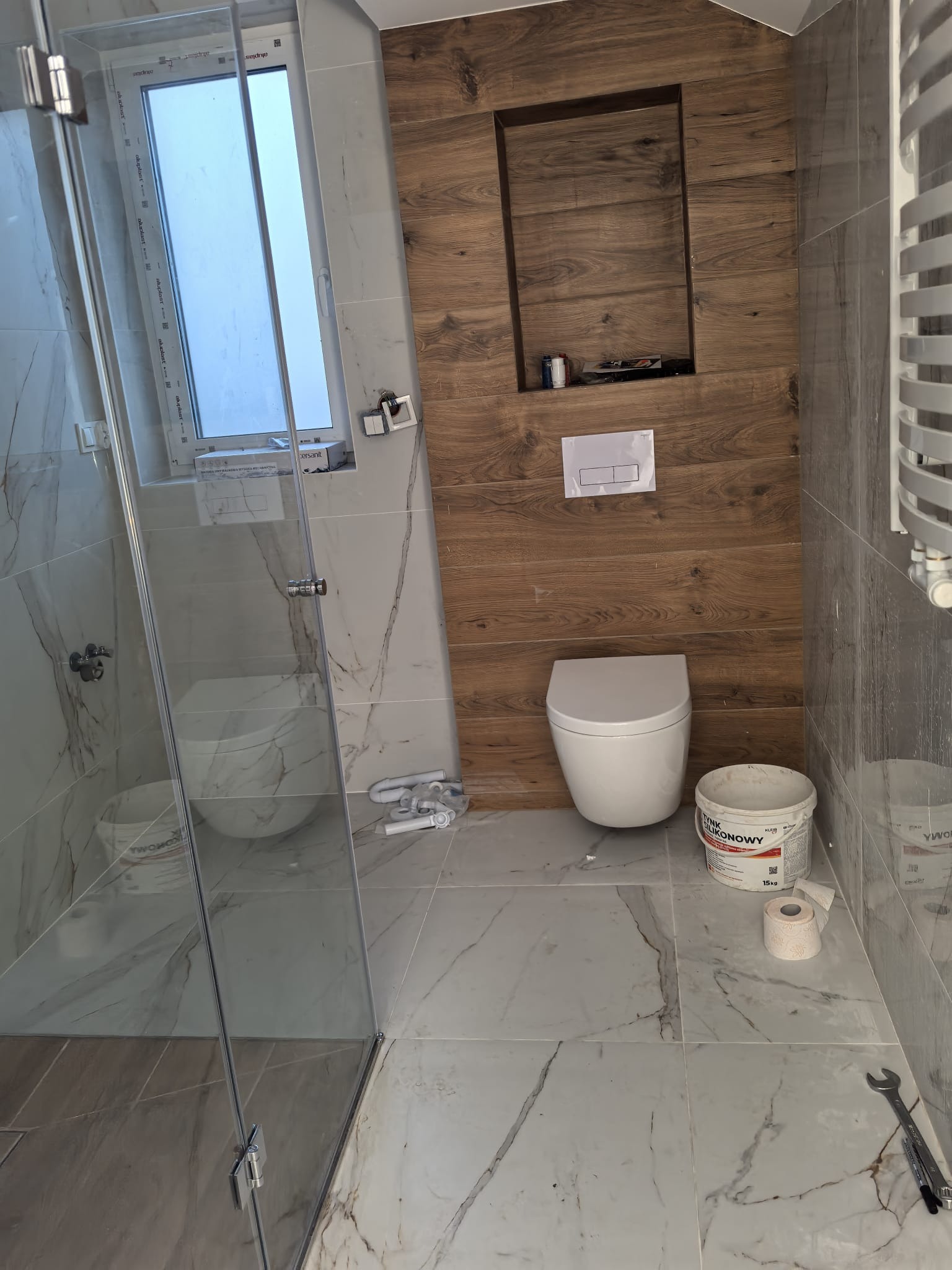 Nowoczesna łazienka w trakcie wykończenia: biała armatura, marmurowe płytki, drewniana ściana z wnęką. Widoczne narzędzia i materiały budowlane.