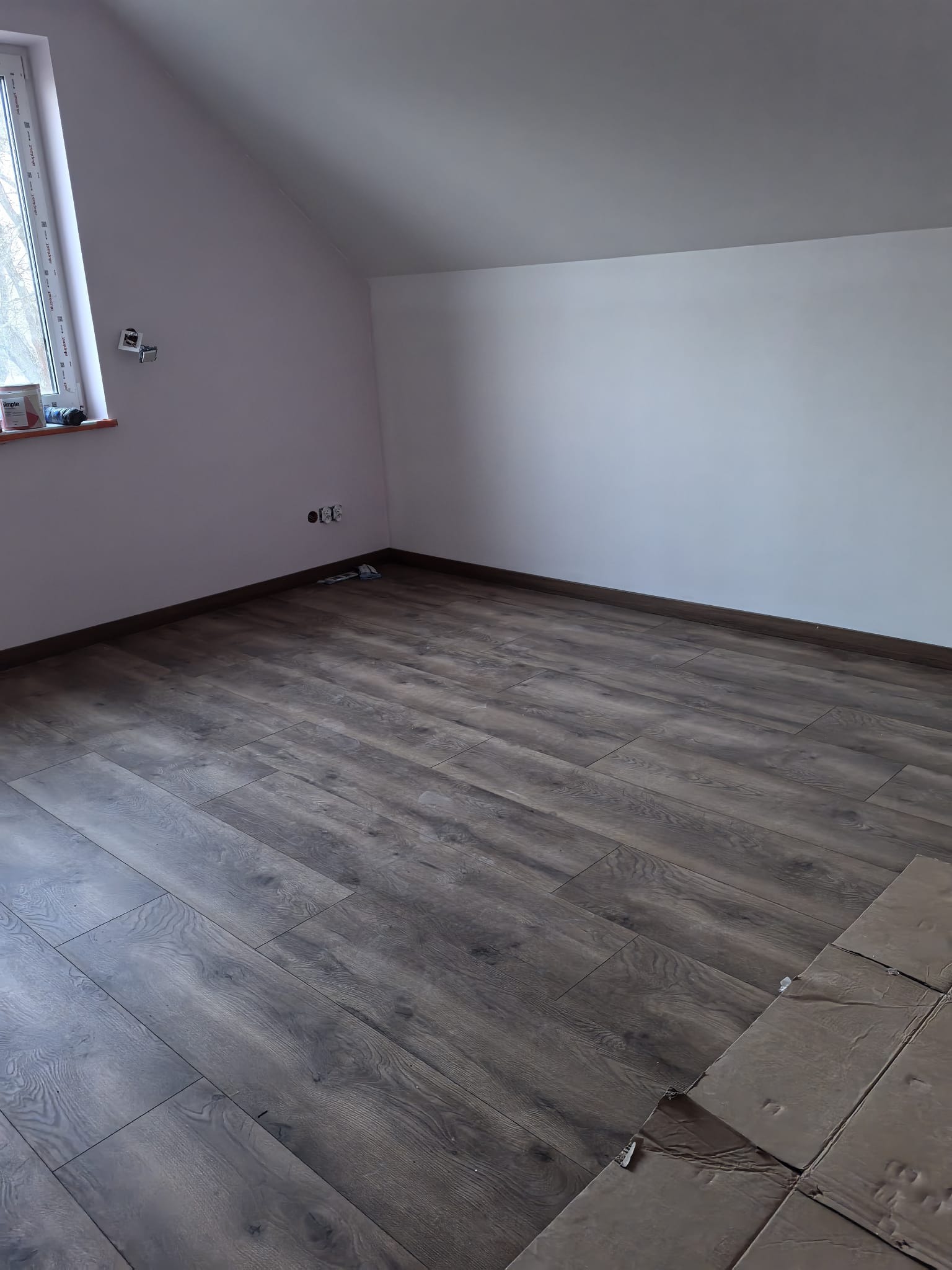 Ukończona podłoga z paneli w pokoju na poddaszu, widoczne fragmenty kartonów zabezpieczających podłogę. Ściany pomalowane na biało i delikatny róż.