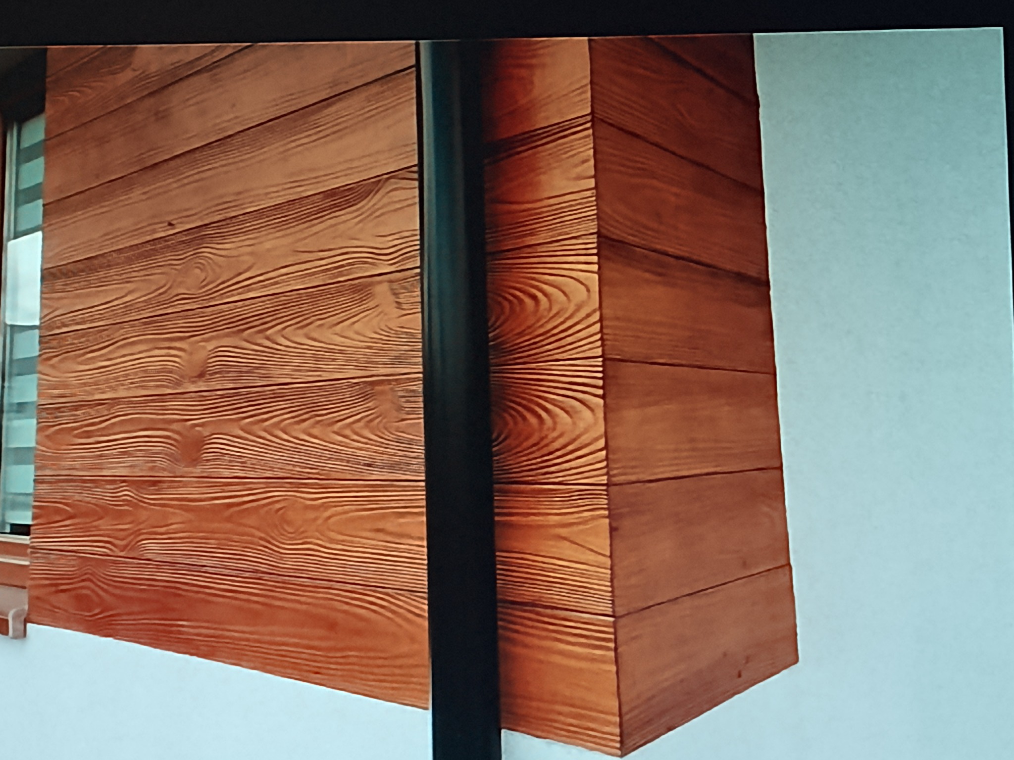 Fragment elewacji budynku z drewnopodobną okładziną, widoczny róg budynku i rura spustowa. Detal wykończenia z bliska, ciepłe odcienie brązu i beżu.