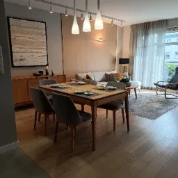 Jasne, przestronne wnętrze mieszkania z drewnianym stołem jadalnym nakrytym dla dwóch osób, szarymi krzesłami, komodą z obrazem, sofą i oświetleniem punktowym oraz trzema wiszącymi lampami.