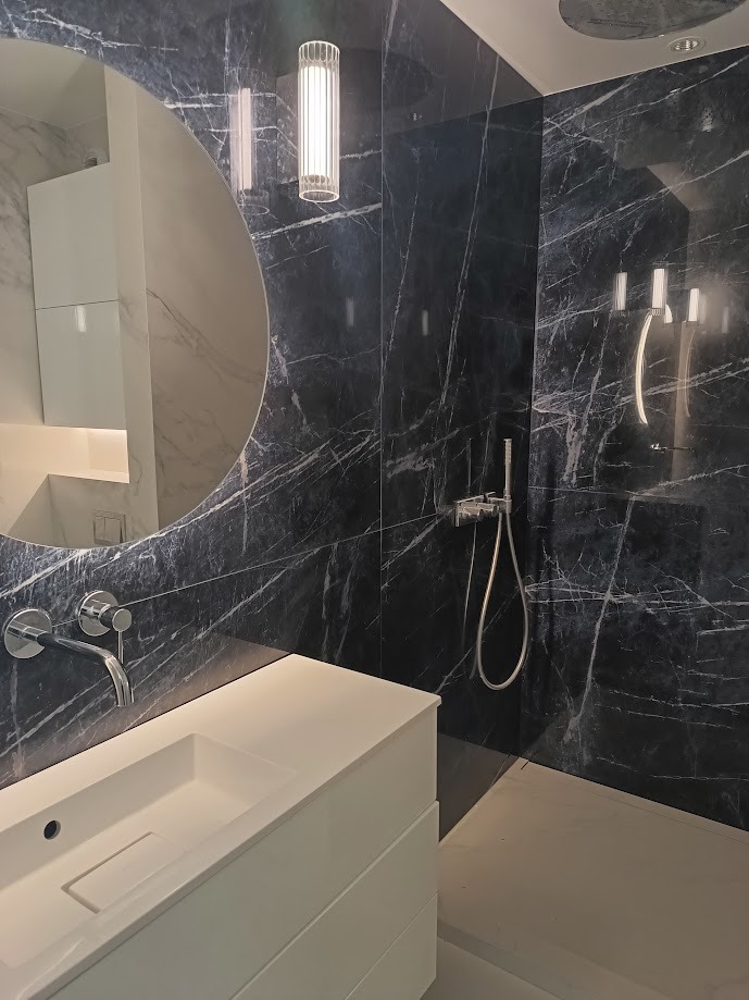 Nowoczesna łazienka z czarnymi marmurowymi ścianami, okrągłym lustrem, białą szafką pod umywalką i chromowaną armaturą.