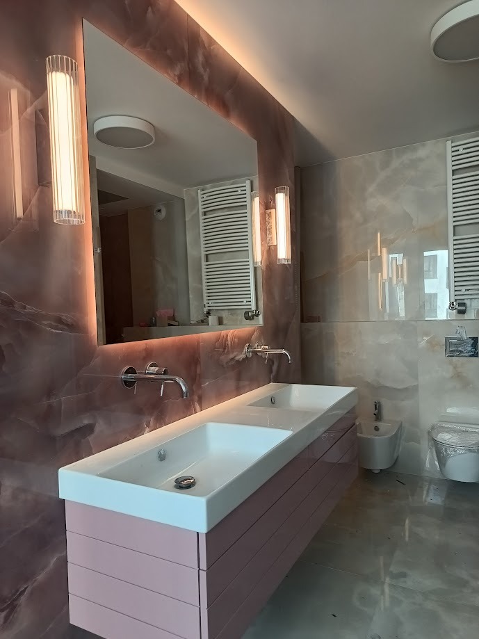 Nowoczesna łazienka z podwójną umywalką nablatową na szafce w odcieniach różu, marmurowe ściany z podświetlanym lustrem i chromowaną armaturą.