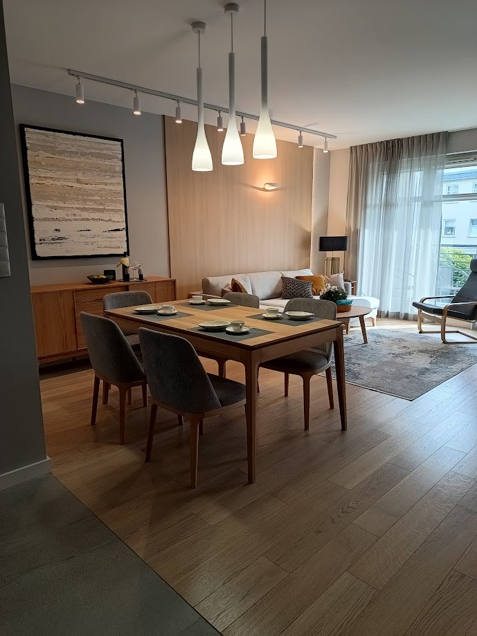 Jasne wnętrze mieszkania z drewnianym stołem jadalnym nakrytym dla czterech osób, szarymi krzesłami, drewnianą komodą z obrazem i sofą w tle, oświetlone lampami wiszącymi i szynoprzewodami.