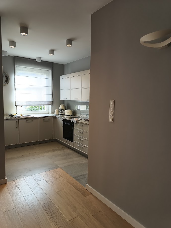 Nowoczesna, jasna kuchnia z drewnianą podłogą, widok z korytarza, z widocznymi sprzętami AGD w stylu retro i minimalistycznym oświetleniem sufitowym.