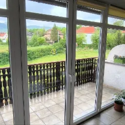 Przesuwne okno balkonowe z białymi ramami, widok na zielony krajobraz z placem zabaw, wewnętrzny balkon z roślinami doniczkowymi i drabinką.