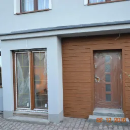 Nowo zamontowane, brązowe drzwi wejściowe z poziomymi panelami, zabezpieczone folią ochronną, obok okno z kratownicą również z folią, na tle jasnoszarej elewacji domu.